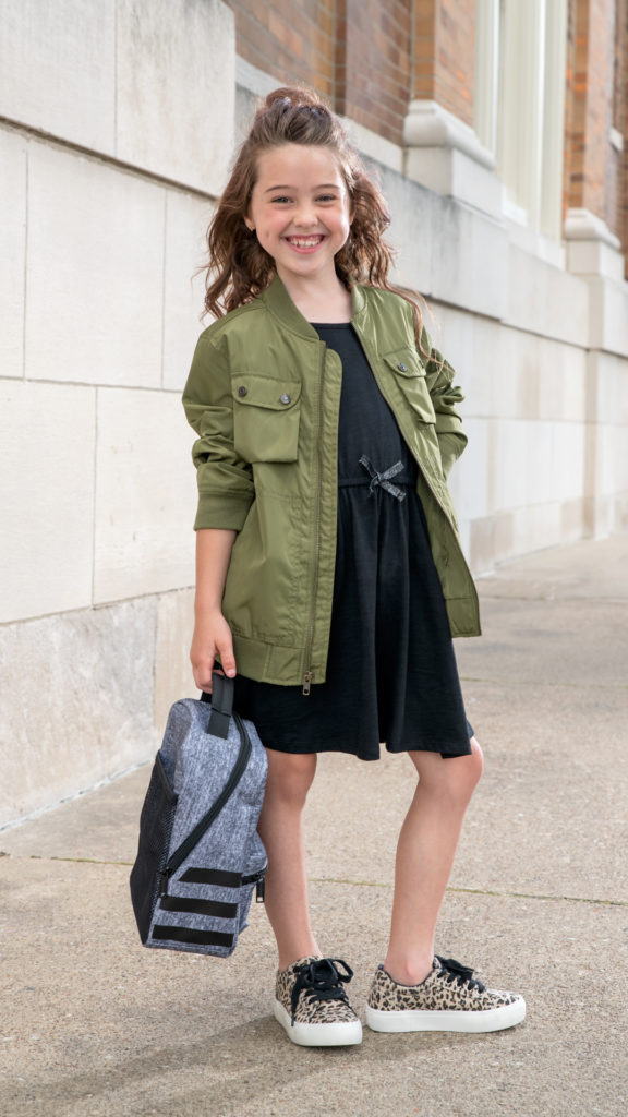 Young girl walking to school and carrying a lunch box. She's wearing a black dress, green jacket, and leopard print sneakers.