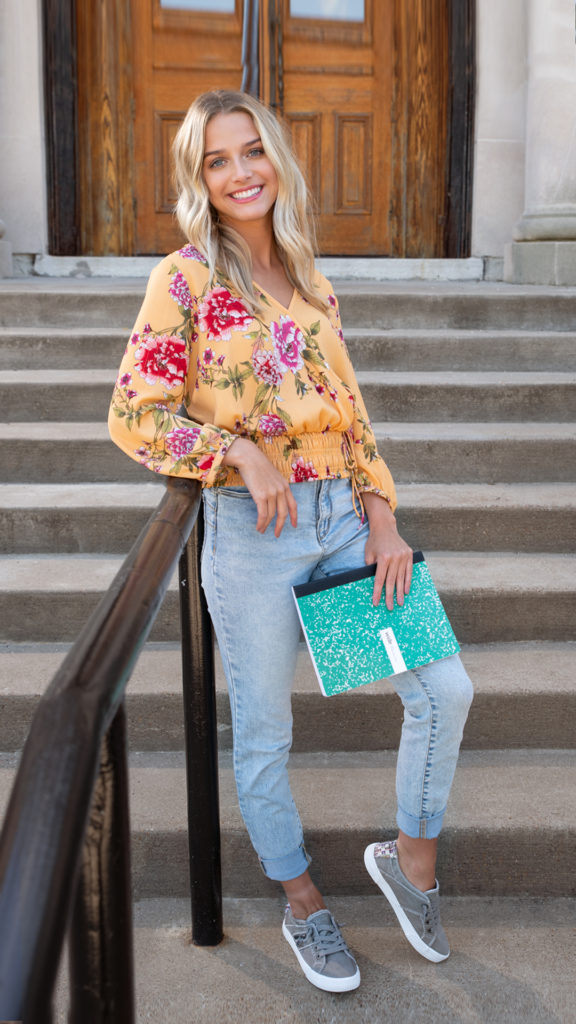 Young woman standing outside the library, wearing a floral blouse and grey sneakers.