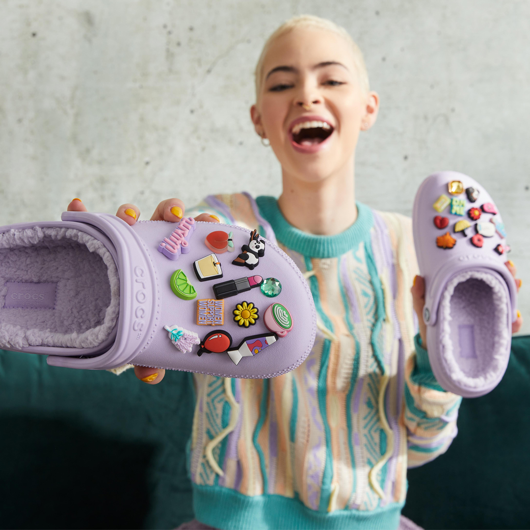 Stylish young person with a big smile shows off fun lined lavender Crocs clogs, which are fully decorated with fun Jibbitz charms