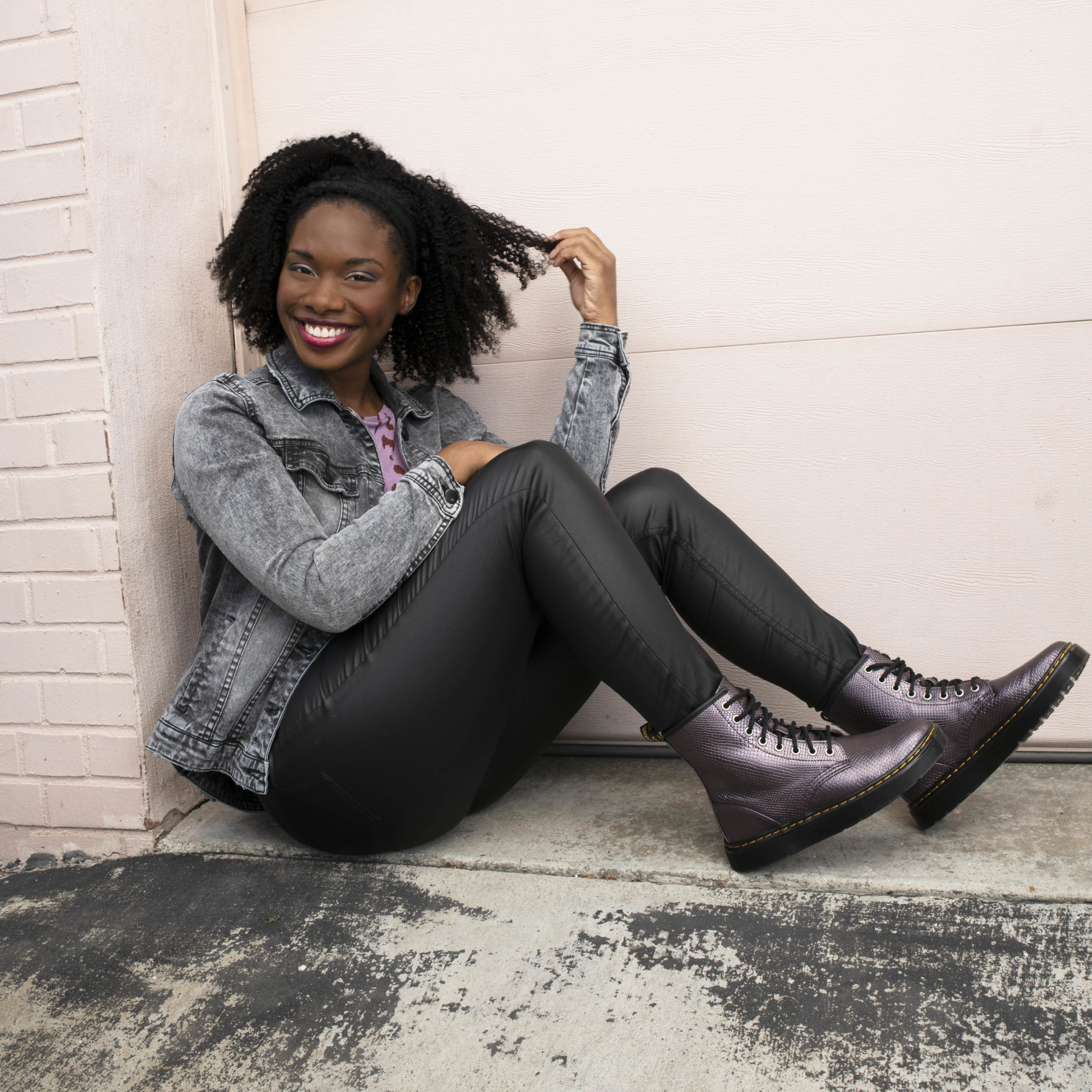 Woman sits in a doorway, twisting her curls and smiling. She's wearing metallic snakeskin combat boots, pleather pants, and a denim jacket. 