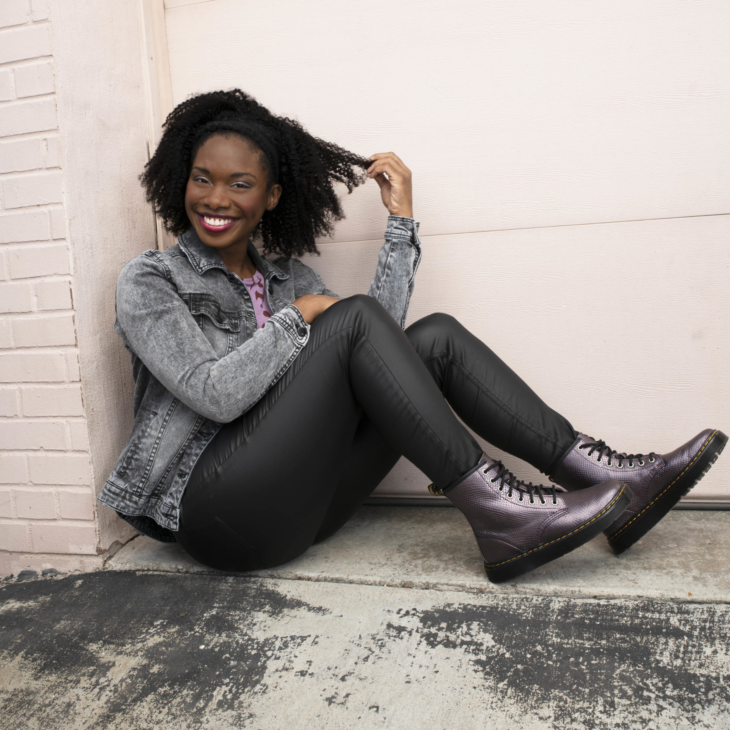 Woman sits in a doorway, twisting her curls and smiling. She's wearing metallic snakeskin combat boots, pleather pants, and a denim jacket.